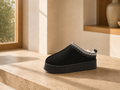 A single black slip-on clog-style shoe with a thick platform sole and a white geometric knit trim around the opening, displayed on a stone ledge in a warm, sunlit interior setting.